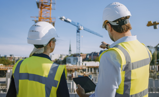 Two people in high-vis vests and white hard hats pointing at a crane on a construction site, holding a tablet