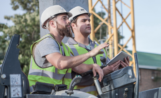 Two men in high-visibility vests on construction machinery at a project site, one holding a tablet